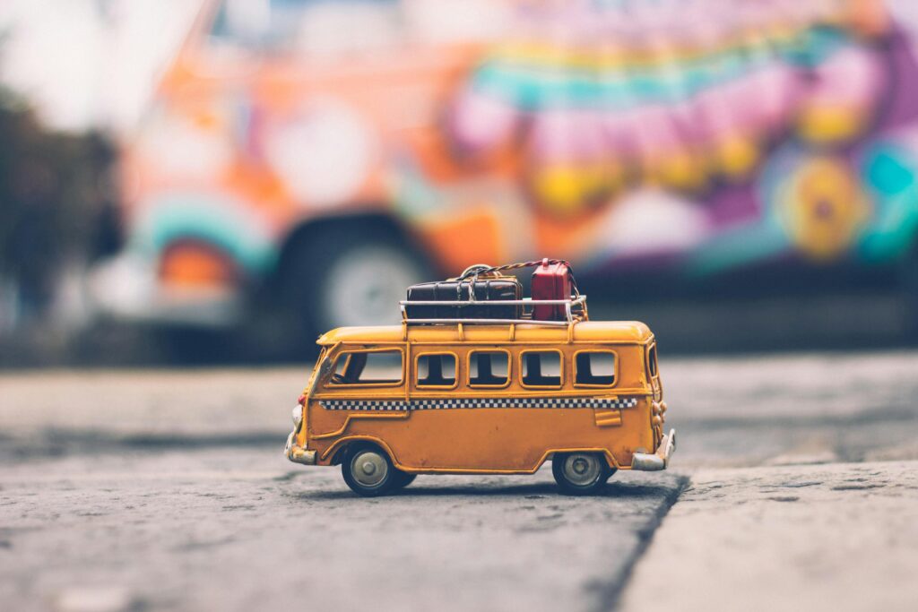 A small toy yellow van with luggage on top is on the ground. In the blurred background, a colorful, graffiti-covered van is visible, creating a playful and adventurous scene.