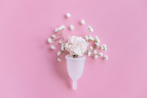 A menstrual cup filled with pink and white flowers, including small blossoms, on a pastel pink background, creating a fresh and gentle tone.